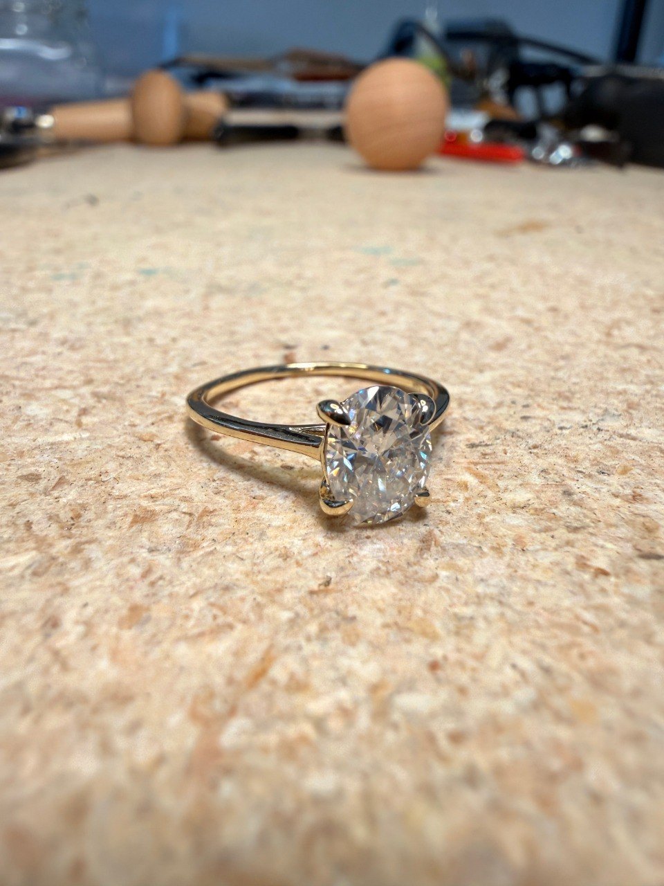 An oval diamond in a yellow gold solitaire setting, sitting on a jeweller's bench next to a ring clamp and hand tools
